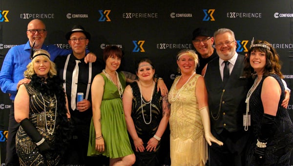 A group in 1920s costumes with pearls, feathers, and suspenders in front of the Experience backdrop