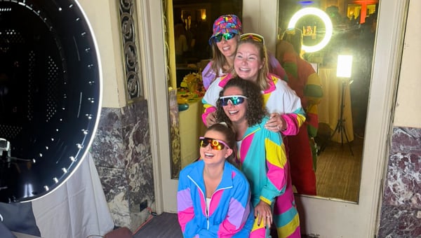 Three attendees in bright 1980s windbreakers and sunglasses posing at a photo booth