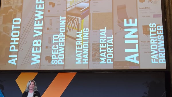 Erin Corrill on stage beneath a screen showing AI Photo, Web Viewer, Spec to PowerPoint, Material Handling, Material Portal, Aline, and Favorites Browser