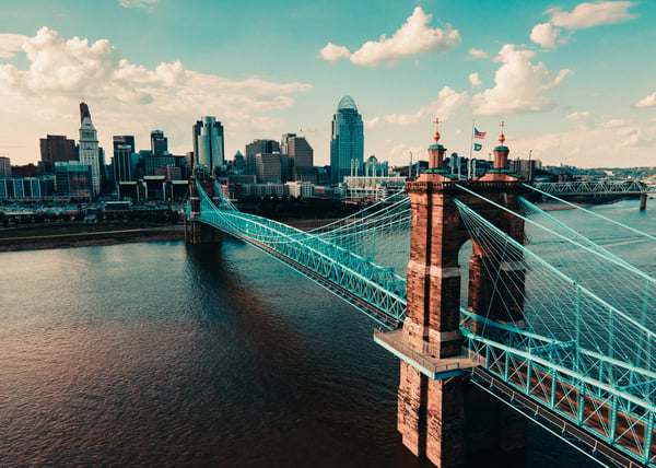 The John A. Roebling Suspension Bridge spanning the Ohio River with the Cincinnati skyline in the background, shown in a vintage-toned photo.